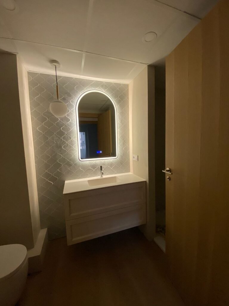 Aseo reformado con azulejo tipo escama, espejo arqueado retroiluminado y mueble de madera a medida