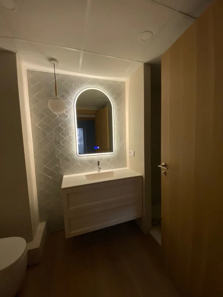 Aseo reformado con azulejo tipo escama, espejo arqueado retroiluminado y mueble de madera a medida