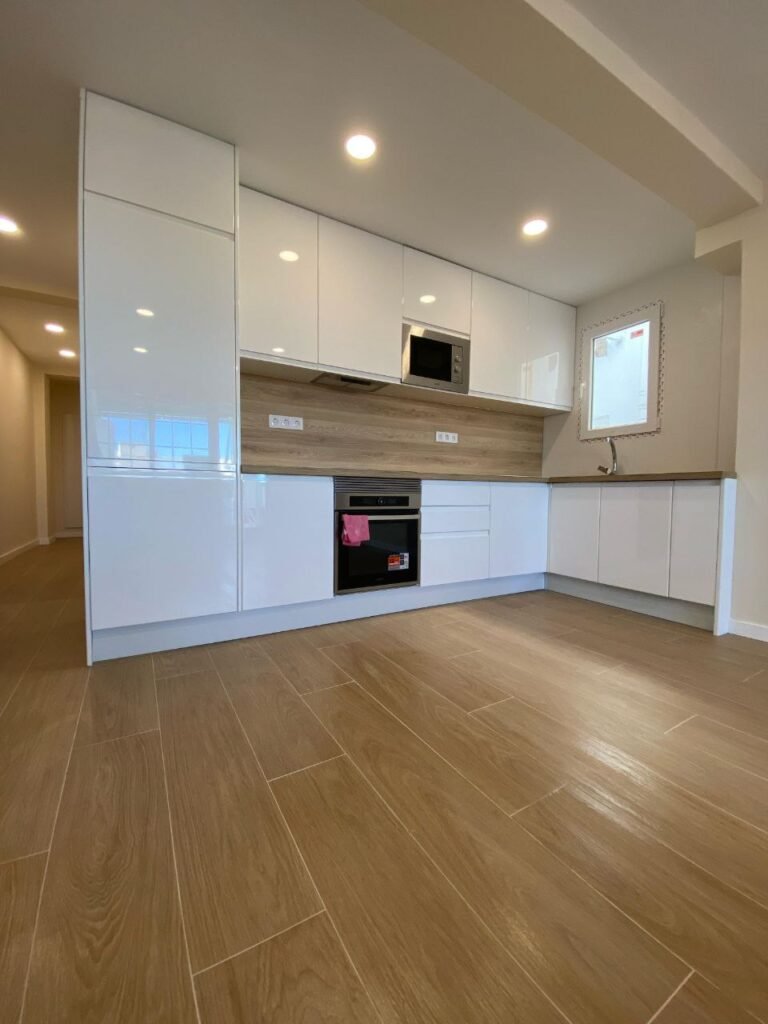Cocina reformada en L con muebles blancos, columna de horno y suelo de tarima
