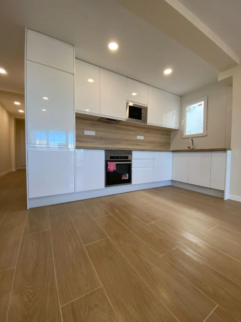 Cocina reformada en L con muebles blancos, columna de horno y suelo de tarima